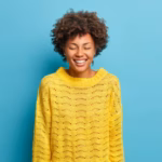 portrait-overjoyed-gentle-woman-smiles-broadly-closes-eyes-shows-white-teeth-wears-casual-yellow-knitted-jumper-expresses-positive-emotions-isolated-blue-wall_273609-44657